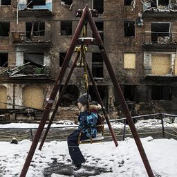 Ein Kind auf einer Schaukel, der Boden ist schneebedeckt, dahinter ein zerstörtes Wohnhaus.