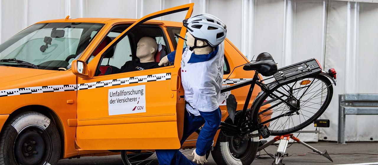 Crashtest-Dummy mit Fahrrad fährt gegen die geöffnete Tür eines geparkten Autos.