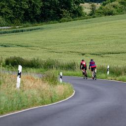 Zwei Rennradfahrer fahren auf einer Landstraße, umgeben von grünen Feldern.