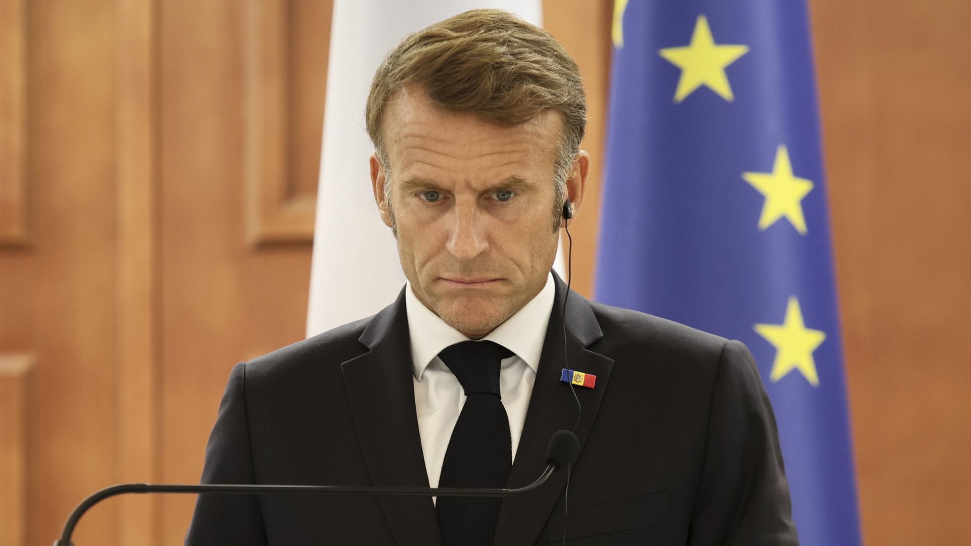Emmanuel Macron bei einer Pressekonferenz. Er hat einen Kopfhörer im Ohr und schaut mit gerunzelter Stirn nach unten. | AP