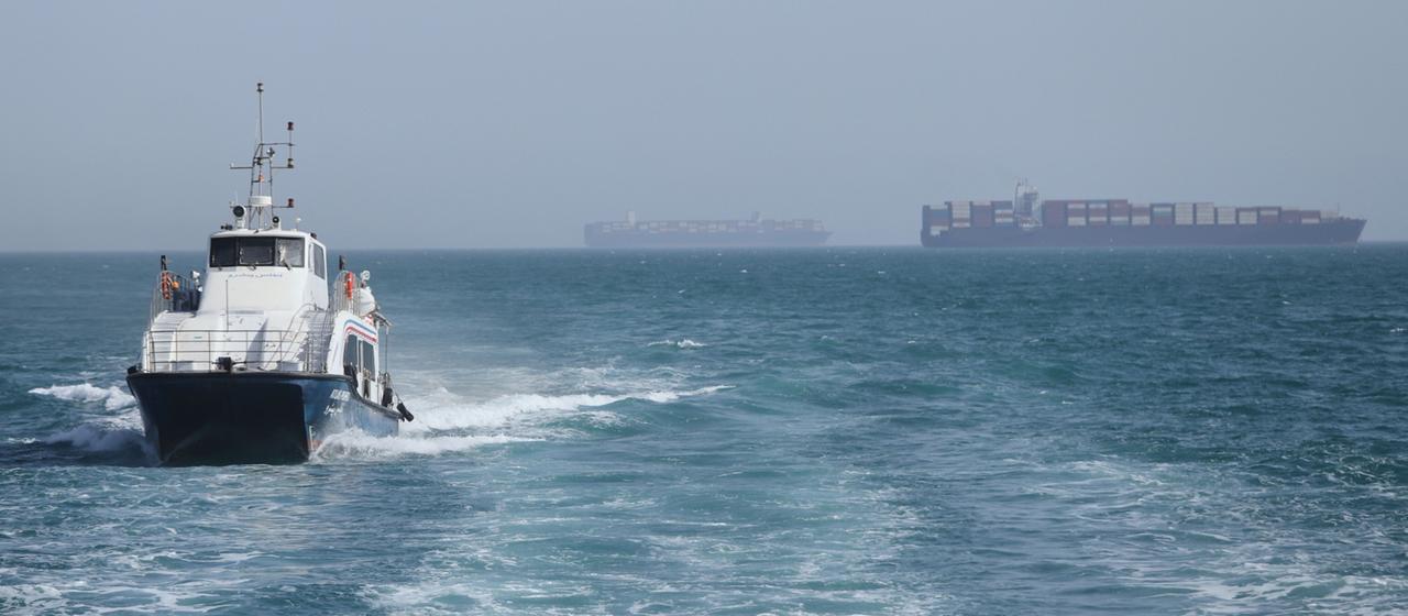  Ein Patrouillenboot fährt durch das Wasser, während Frachtschiffe in der Straße von Hormus vor Anker liegen. 